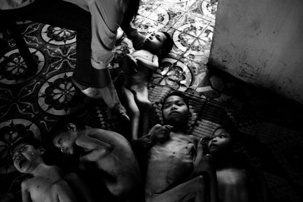 Group of bare-chested children lying on a patterned tiled floor, looking up with tired expressions as a caregiver reaches toward them.