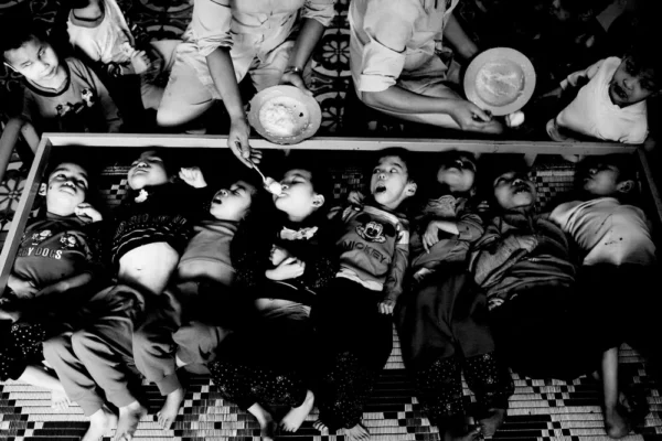Black-and-white photo of many children lying side by side in a trough, mouths open as caregivers feed them with spoons from above.