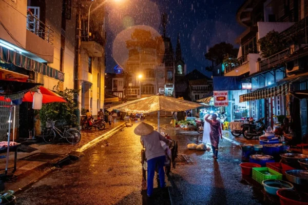 Night street market in heavy rain; a vendor with a conical hat pushes a cart under a beige umbrella while the wet street reflects lights at night.