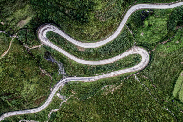 Aerial view of a winding white road cutting through dense green hillside vegetation.