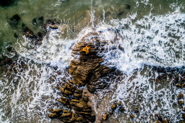 Aerial view of a person in an orange jacket standing on jagged rocks as waves crash around them at the shoreline