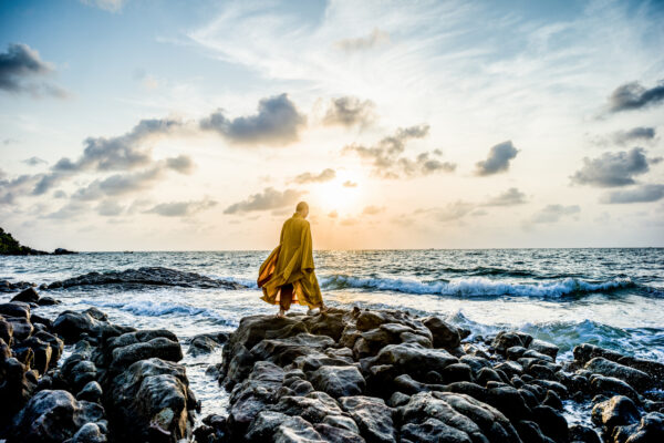 Person wearing a yellow cloak walking on rocky shoreline at sunset by the ocean