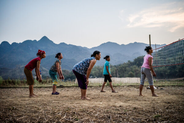 Women in colorful outfits bending forward to play volleyball in an open field, with a net on the right and mountains in the distance.